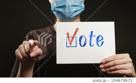 Vote sign. Pandemic agitation. Your choice. Unrecognizable woman in face mask showing bulletin with check mark pointing finger on dark. Vote sign. Pandemic agitation. Your choice. Unrecognizable woman in face mask showing bulletin with check mark pointing finger on dark. 104678672