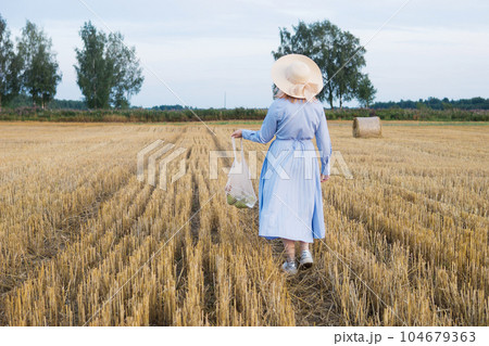 A red-haired woman in a hat and a blue dress walks in a field with haystacks. A red-haired woman in a hat and a blue dress walks in a field with haystacks. 104679363