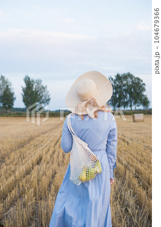 A red-haired woman in a hat and a blue dress walks in a field with haystacks. 104679366