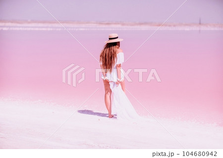Woman in pink salt lake. She in a white dress and hat enjoys the scenic view of a pink salt lake as she walks along the white, salty shore, creating a lasting memory. 104680942