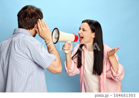 Woman shouting in megaphone to man against blue studio background. Emotional conversation and misunderstanding 104681303