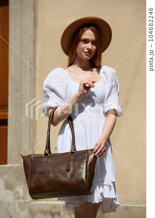young beautiful redhair girl with red lipstick in a big hat and white summer dress with brown handbag posing on the streets young beautiful redhair girl with red lipstick in a big hat and white summer dress with brown handbag posing on the streets 104682246
