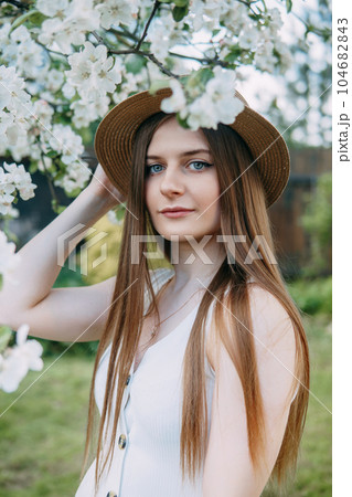 Beautiful young girl in white dress and hat in blooming Apple orchard. Blooming Apple trees with white flowers. Beautiful young girl in white dress and hat in blooming Apple orchard. Blooming Apple trees with white flowers. 104682843