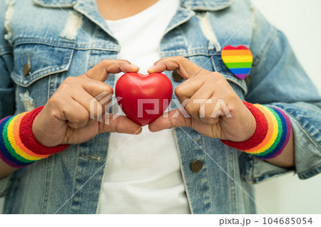 Asian lady wearing rainbow flag wristbands and hold red heart, symbol of LGBT pride month celebrate annual in June social of gay, lesbian, bisexual, transgender, human rights. 104685054