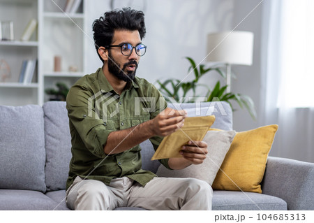 Upset and sad man at home received notification mail envelope with bad news, hindu man sitting on sofa in living room depressed reading and holding envelope with message in hands. 104685313