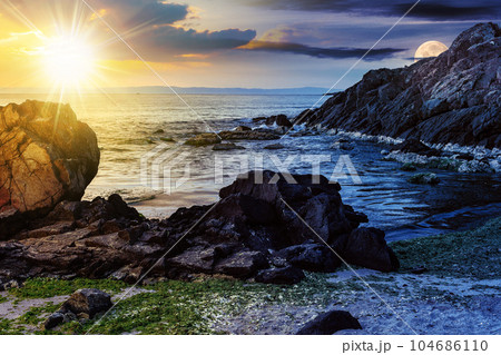 day and night time change concept. number of stones and rock on the sandy sea coast with sun and moon at twilight. seaweed lie on the sand made by storm in morning light 104686110