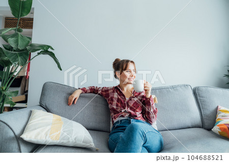 Young smiling woman sitting on sofa and looking away while drinking coffee or tea. Young brunette woman relaxing after housekeeping, home cleaning. Portrait of relaxed female resting at home. Young smiling woman sitting on sofa and looking away while drinking coffee or tea. Young brunette woman relaxing after housekeeping, home cleaning. Portrait of relaxed female resting at home. 104688521
