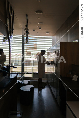 Vertical silhouette shot of unrecognizable man in white robe after spa treatment at wellness center opening locker to change clothes in dark dressing room with mirrored walls, standing by window. Vertical silhouette shot of unrecognizable man in white robe after spa treatment at wellness center opening locker to change clothes in dark dressing room with mirrored walls, standing by window. 104688524