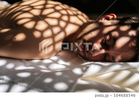 Close up face of sleeping handsome man relaxing lying down with closed eyes on massage table in sunlight and beautiful shadows in spa salon. Concept of body care and rehabilitation of spiritual peace 104688546