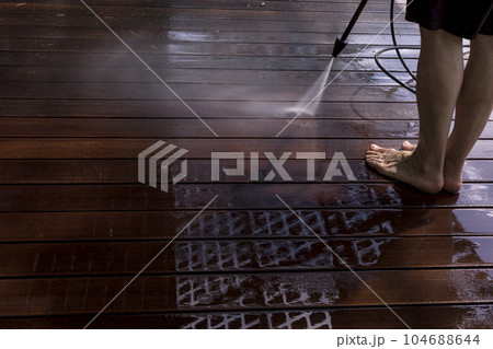 Power surface washing. Man cleaning terrace with a power washer. High water pressure cleaner on wooden terrace surface. 104688644