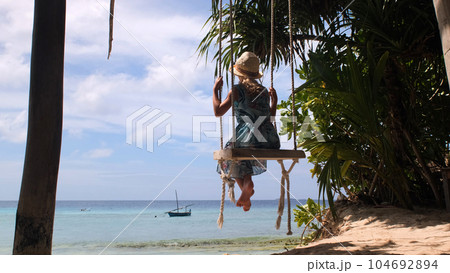 Little girl in straw hat and blue sundress enjoying ocean landscape while swinging on tropical beach 104692894