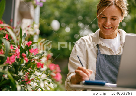 Close up of flower shop owner wearing apron working on laptop and taking online orders in store Close up of flower shop owner wearing apron working on laptop and taking online orders in store 104693863
