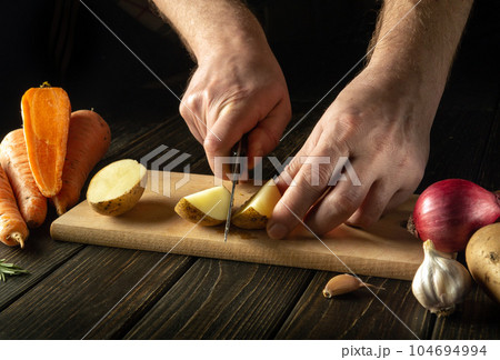 Cutting raw potatoes on a wooden cutting board with a knife in the hands of a chef. Cooking a vegetable dish in a restaurant kitchen 104694994