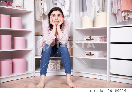 A young beautiful brunette woman in jeans and a pink jacket sits in a dressing room with her face in her hands meditating A young beautiful brunette woman in jeans and a pink jacket sits in a dressing room with her face in her hands meditating 104696144