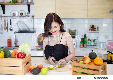 beautiful asian woman smiling preparing, cut green apple in white kitchen with plenty of fresh vegetables and fruits on the table. 104699661