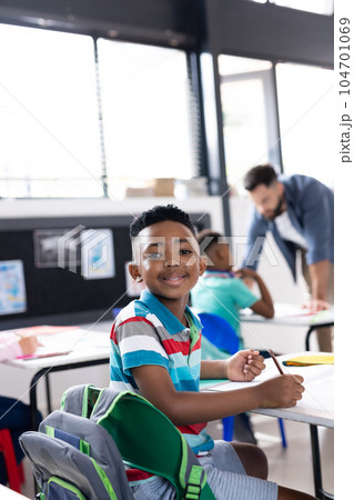 Vertical portrait of smiling african american schoolboy at desk in elementary classroom, copy space 104701069