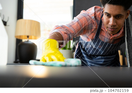 Focused biracial man wearing apron and rubber gloves cleaning countertop in sunny kitchen 104702159