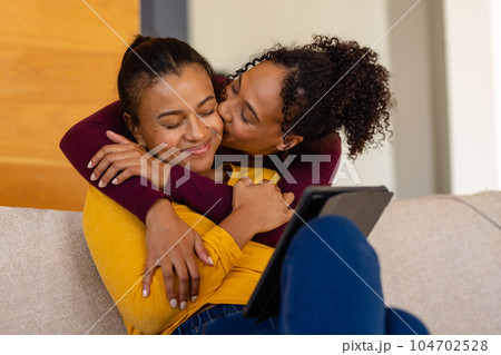 Happy biracial lesbian couple hugging and using tablet on sofa in living room 104702528