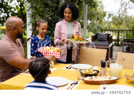 Happy biracial parents, son and daughter serving food and sitting at table for meal on terrace 104702759