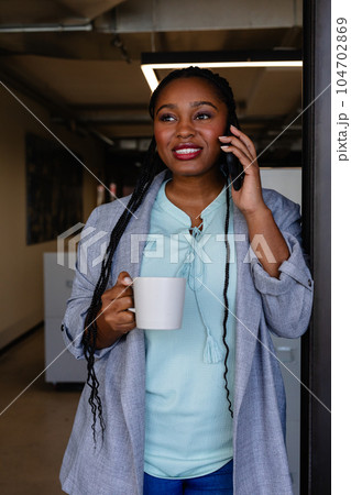 Plus size african american casual businesswoman with cup of coffee talking on smartphone in office 104702869