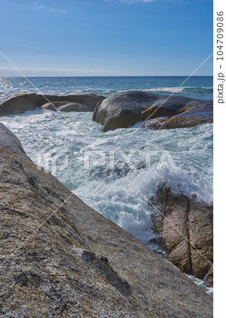 Landscape view of boulders on the ocean and water during summer. Scenic view of waves crashing into the rocks in the sea against a clear blue sky. Beautiful nature views at the beach on the coast 104709086