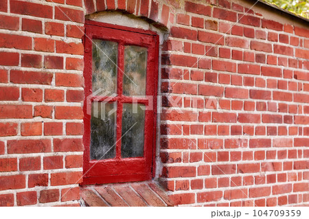Old dirty window in a red brick house or home. Ancient casement with red wood frame on a historic building with clumpy paint texture. Exterior details of a windowsill in a traditional town or village 104709359