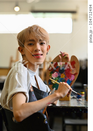 Cheerful young man art school student sitting in workshop holding palette and paintbrush, smiling at camera 104712494