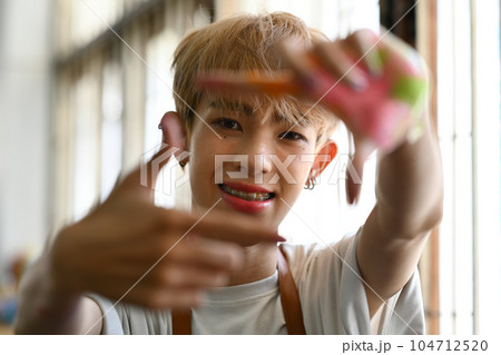 Happy young queer man framing his face with hands, looking at camera with a smile while standing in art studio Happy young queer man framing his face with hands, looking at camera with a smile while standing in art studio 104712520