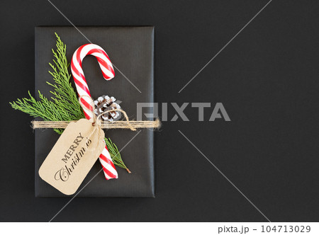 Black gift box with candy cane, cedar tree sprig, cone and label with the text merry christmas on dark background. Space for yout text. Black gift box with candy cane, cedar tree sprig, cone and label with the text merry christmas on dark background. Space for yout text. 104713029