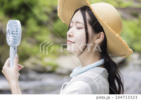 川辺でネッククーラーをして携帯扇風機を使う麦わら帽子を被った若い女性 104713323