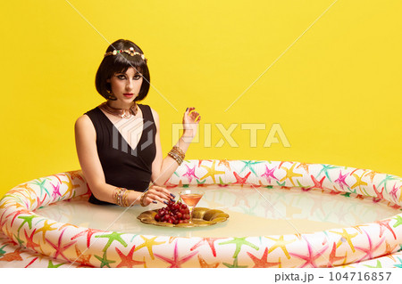 Beautiful young woman in image of Cleopatra sitting in swimming pool with milk and eating breakfast against yellow studio background. Beautiful young woman in image of Cleopatra sitting in swimming pool with milk and eating breakfast against yellow studio background. 104716857