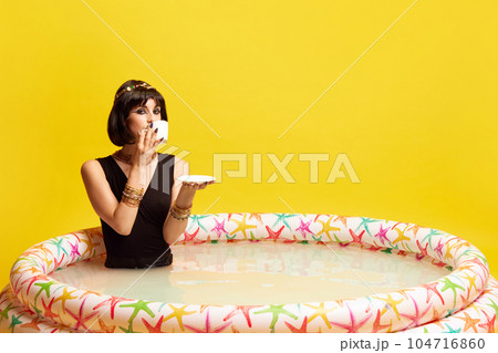 Pretty young woman in image of Cleopatra sitting in swimming pool with milk and drinking coffe against yellow studio background. Pretty young woman in image of Cleopatra sitting in swimming pool with milk and drinking coffe against yellow studio background. 104716860