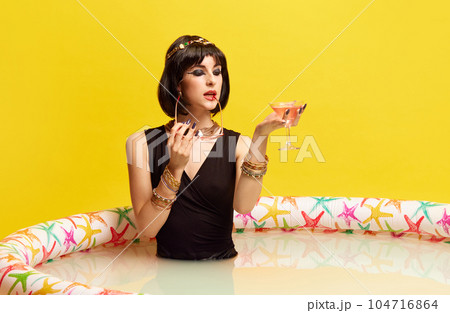 Party. Pretty young woman in image of Cleopatra sitting in swimming pool with milk and drinking cocktail against yellow studio background. 104716864