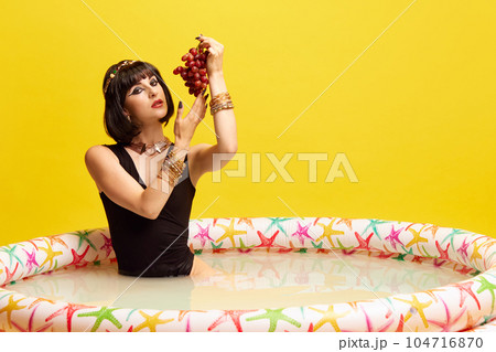 Beautiful young woman in image of Cleopatra sitting in swimming pool with milk and eating grapes against yellow studio background. 104716870