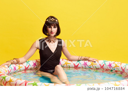 Portrait of attractive, beautiful young woman with expressive makeup, in image of Cleopatra sitting in swimming pool against yellow studio background 104716884