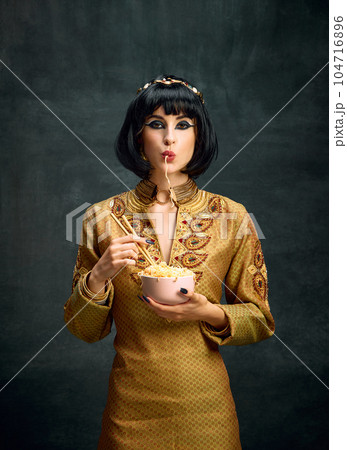 Tasty. Beautiful young woman in image of egyptian queen, Cleopatra eating instant noodles against dark vintage background Tasty. Beautiful young woman in image of egyptian queen, Cleopatra eating instant noodles against dark vintage background 104716896