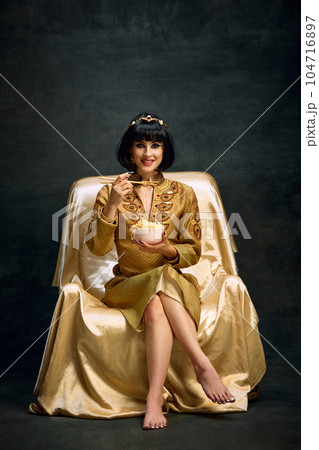 Portrait of smiling young woman in image of egyptian queen, Cleopatra eating instant noodles against dark vintage background Portrait of smiling young woman in image of egyptian queen, Cleopatra eating instant noodles against dark vintage background 104716897