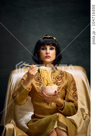 Portrait of beautiful young woman in image of egyptian queen, Cleopatra eating instant noodles against dark vintage background 104716898