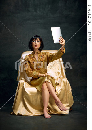 Portrait of young beautiful woman, queen, Cleopatra in golden dress taking selfie with tablet against dark vintage background Portrait of young beautiful woman, queen, Cleopatra in golden dress taking selfie with tablet against dark vintage background 104716911