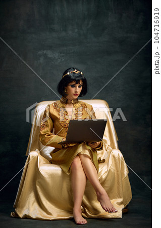 Portrait of beautiful young woman in image of queen, Cleopatra sitting in golden dress and working on laptop against dark vintage background Portrait of beautiful young woman in image of queen, Cleopatra sitting in golden dress and working on laptop against dark vintage background 104716919