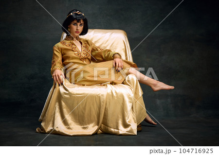 Portrait of elegant, beautiful woman in image of antique queen, Cleopatra in olden clothes sitting on armchair against dark vintage background Portrait of elegant, beautiful woman in image of antique queen, Cleopatra in olden clothes sitting on armchair against dark vintage background 104716925