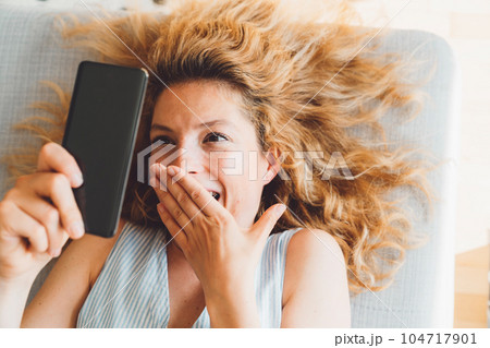 Playful woman taking funny selfies while laying down on the couch with her hair spread out on the surface Playful woman taking funny selfies while laying down on the couch with her hair spread out on the surface 104717901