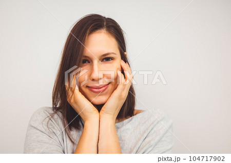 Portrait of young woman holding hands on her face with a sbtle smile looking at the camera on white background Portrait of young woman holding hands on her face with a sbtle smile looking at the camera on white background 104717902