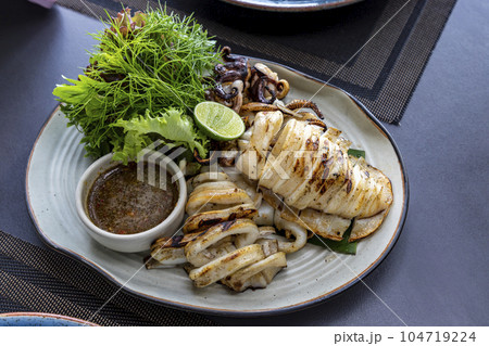 A large grilled squid looks appetizing with a small cup of spicy sauce next to it, served with vegetables in a white porcelain plate. On a table in a restaurant. Selective focus. 104719224