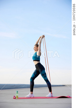 Feeling positive. Young sportive woman in stylish sportswear training fitn fitness elastic bands outdoors on warm sunny day 104720086