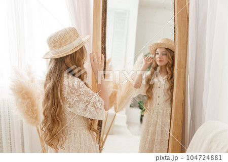 A beautiful teenage girl with long hair measures a straw hat in front of a mirror. Self-admiration of a blonde. 104724881