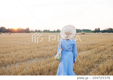A red-haired woman in a hat and a blue dress walks in a field with haystacks. 104725376