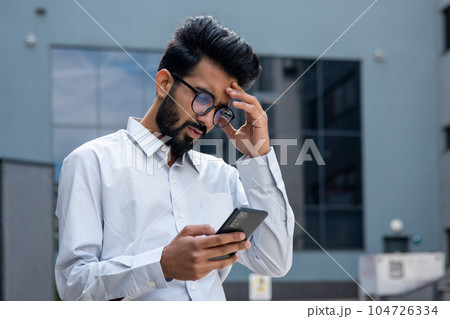 Young hispanic businessman outside office building, man in shirt reading sad bad news online using app on phone, depressed man disappointed unhappy with achievement results. Young hispanic businessman outside office building, man in shirt reading sad bad news online using app on phone, depressed man disappointed unhappy with achievement results. 104726334
