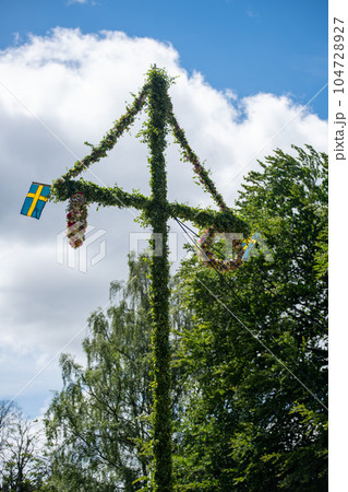 A pole and flag against green trees and blue sky. A maypole decorated, covered in flowers and leaves. A pole and flag against green trees and blue sky. A maypole decorated, covered in flowers and leaves. 104728927