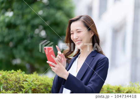 屋外にてスマホの画面を見て笑顔になるビジネスウーマン 屋外にてスマホの画面を見て笑顔になるビジネスウーマン 104734418